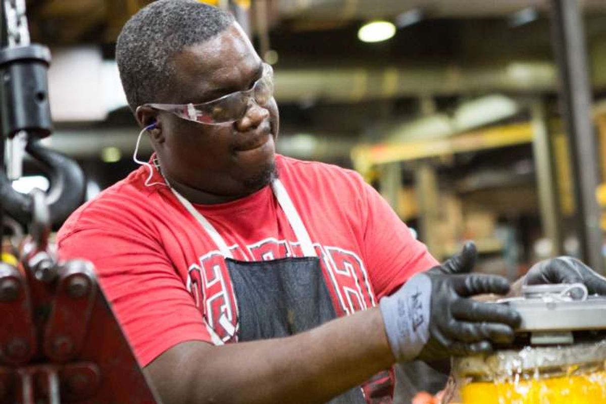 A man wearing safety glasses, gloves, and a red shirt works with machinery in an industrial setting. The background is blurred with equipment visible.