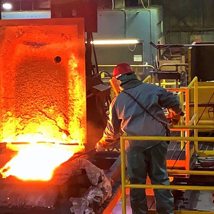 A worker in protective gear stands near molten metal pouring from a large furnace in an industrial foundry setting.