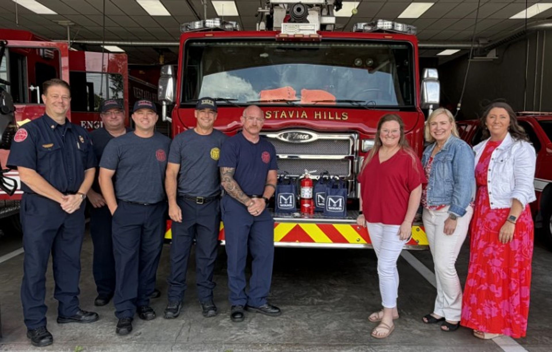 Seven people stand smiling in front of a firetruck inside a fire station, with two bags and drinks placed on the truck’s bumper. Four are in firefighter uniforms, three are in casual clothes.