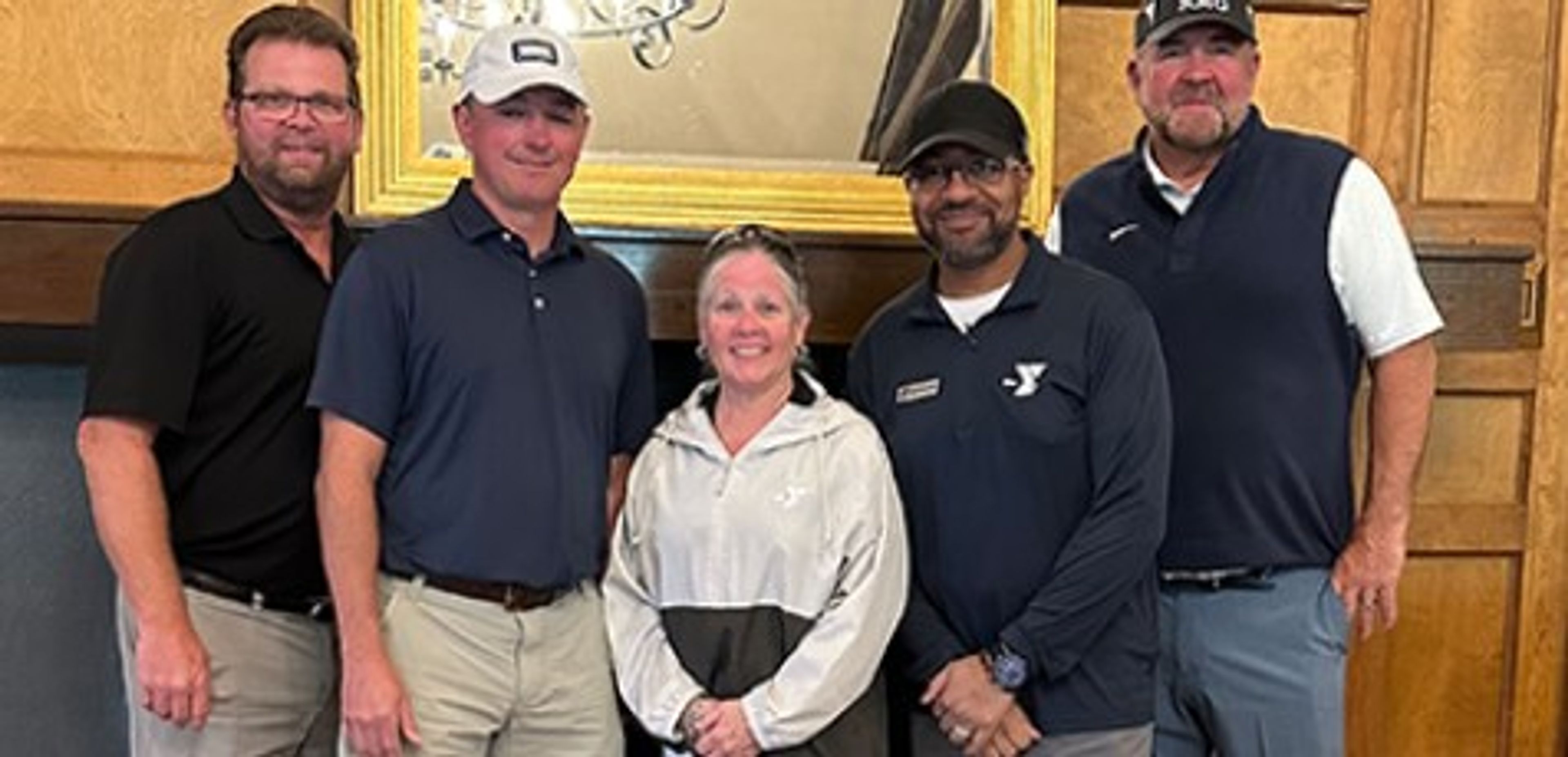 Five adults pose indoors in front of a wooden fireplace and mirror. Four men in collared shirts and hats stand beside a woman in a light jacket. All are smiling at the camera.