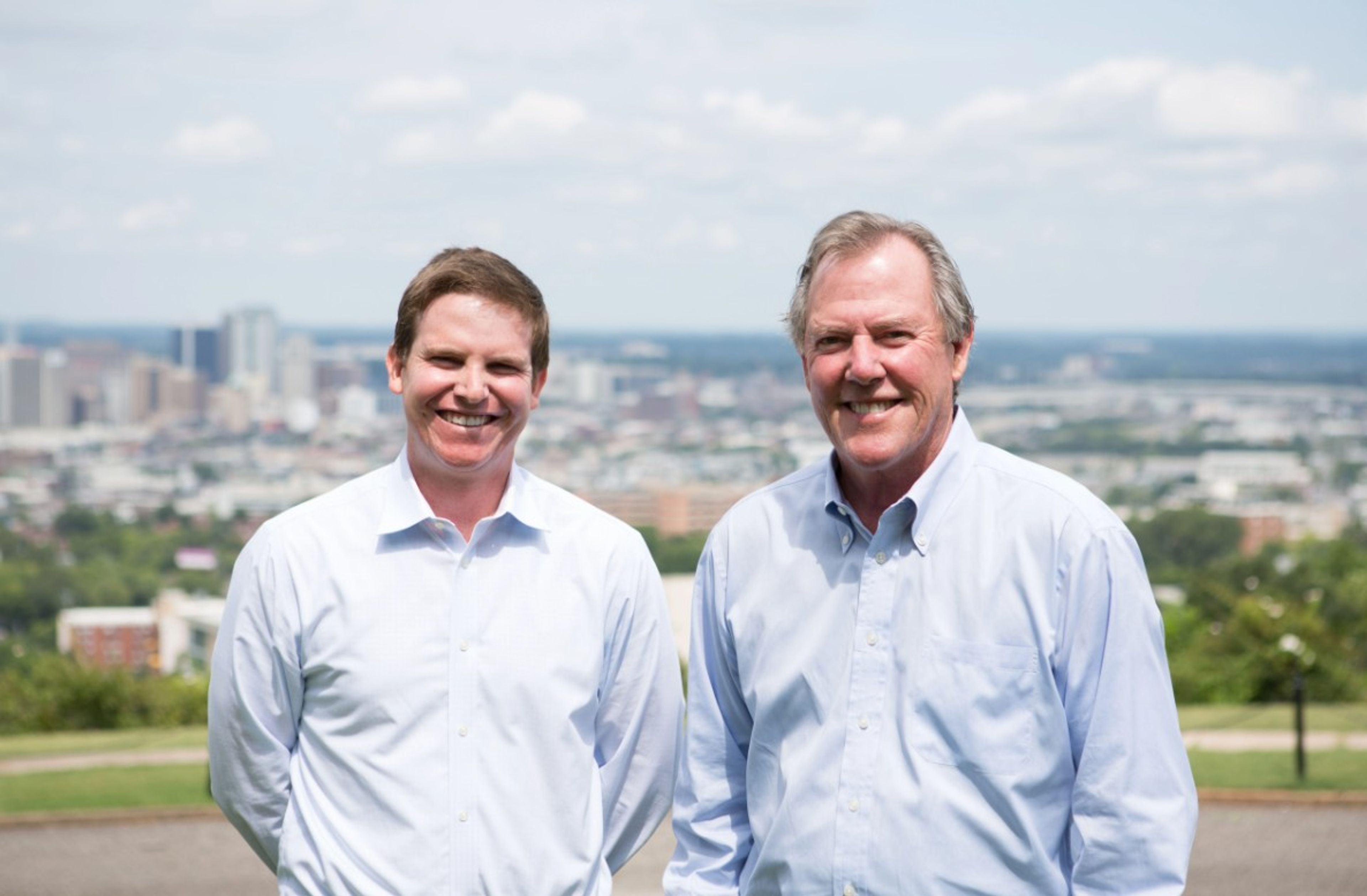 Two men in light blue shirts stand outside, smiling at the camera, with a cityscape and cloudy sky in the background.