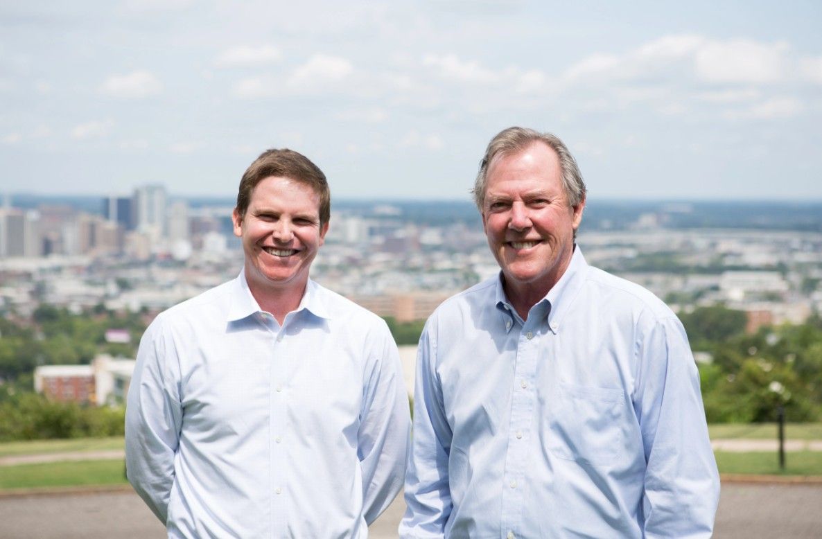 Two men in light blue shirts stand outside, smiling at the camera, with a cityscape and cloudy sky in the background.