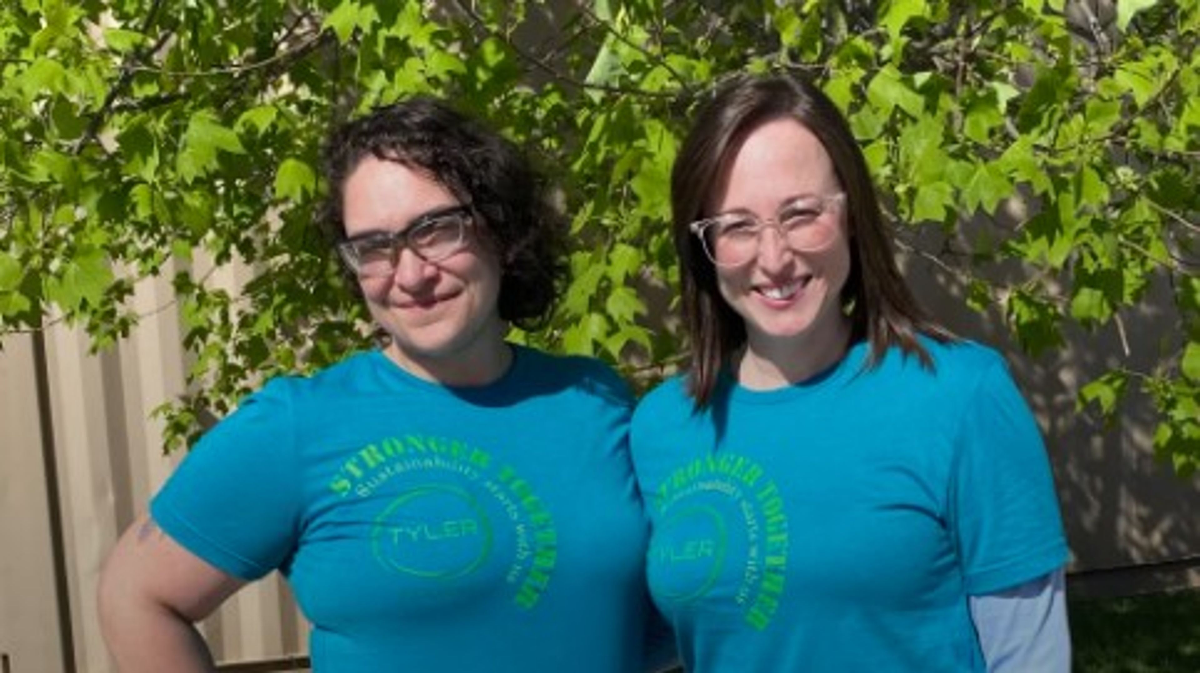 Two women wearing matching blue “Stronger Together” T-shirts and glasses stand outside in front of leafy green trees, smiling at the camera in bright sunlight.