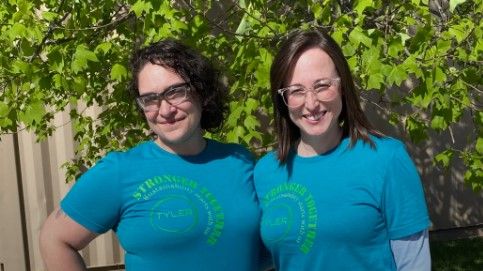 Two women wearing matching blue “Stronger Together” T-shirts and glasses stand outside in front of leafy green trees, smiling at the camera in bright sunlight.