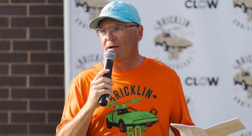 A man in an orange “Bricklin 50th” shirt and blue cap speaks into a microphone, holding papers. A Bricklin event banner is visible behind him.
