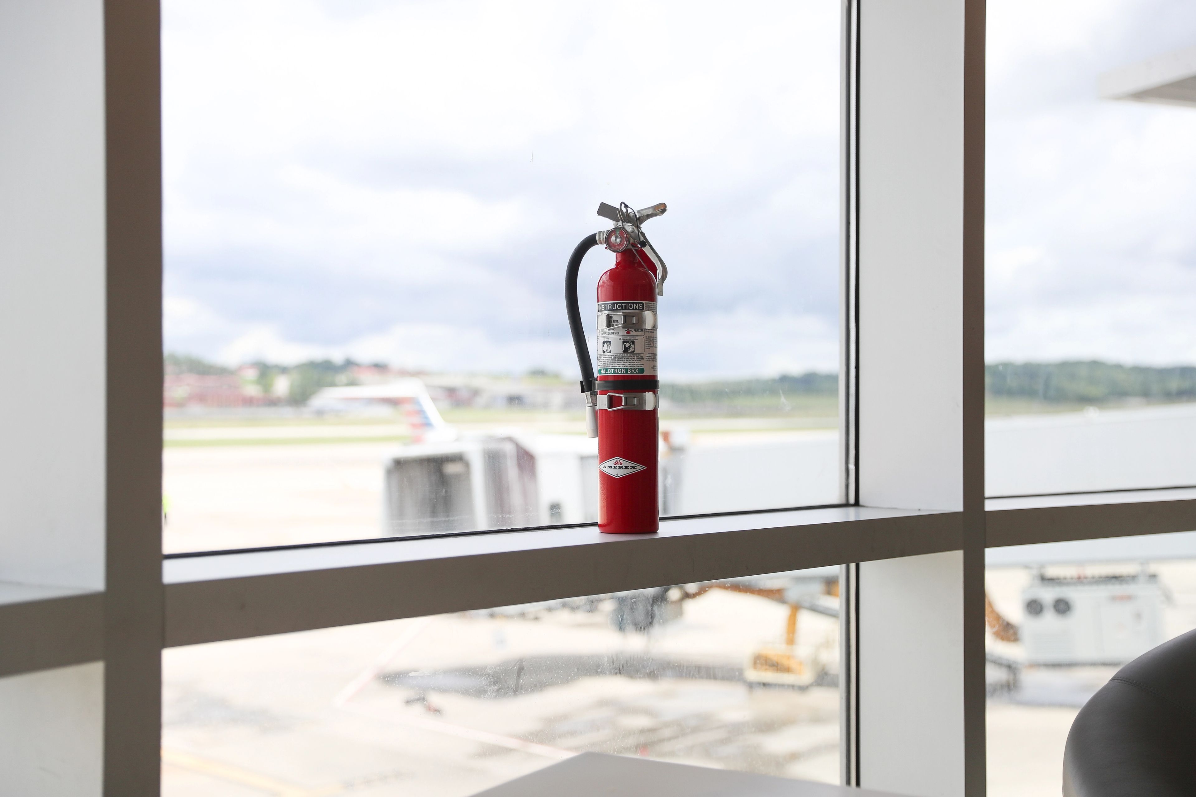 A red fire extinguisher sits on a windowsill at an airport, with airplanes and a runway visible through the large window in the background.