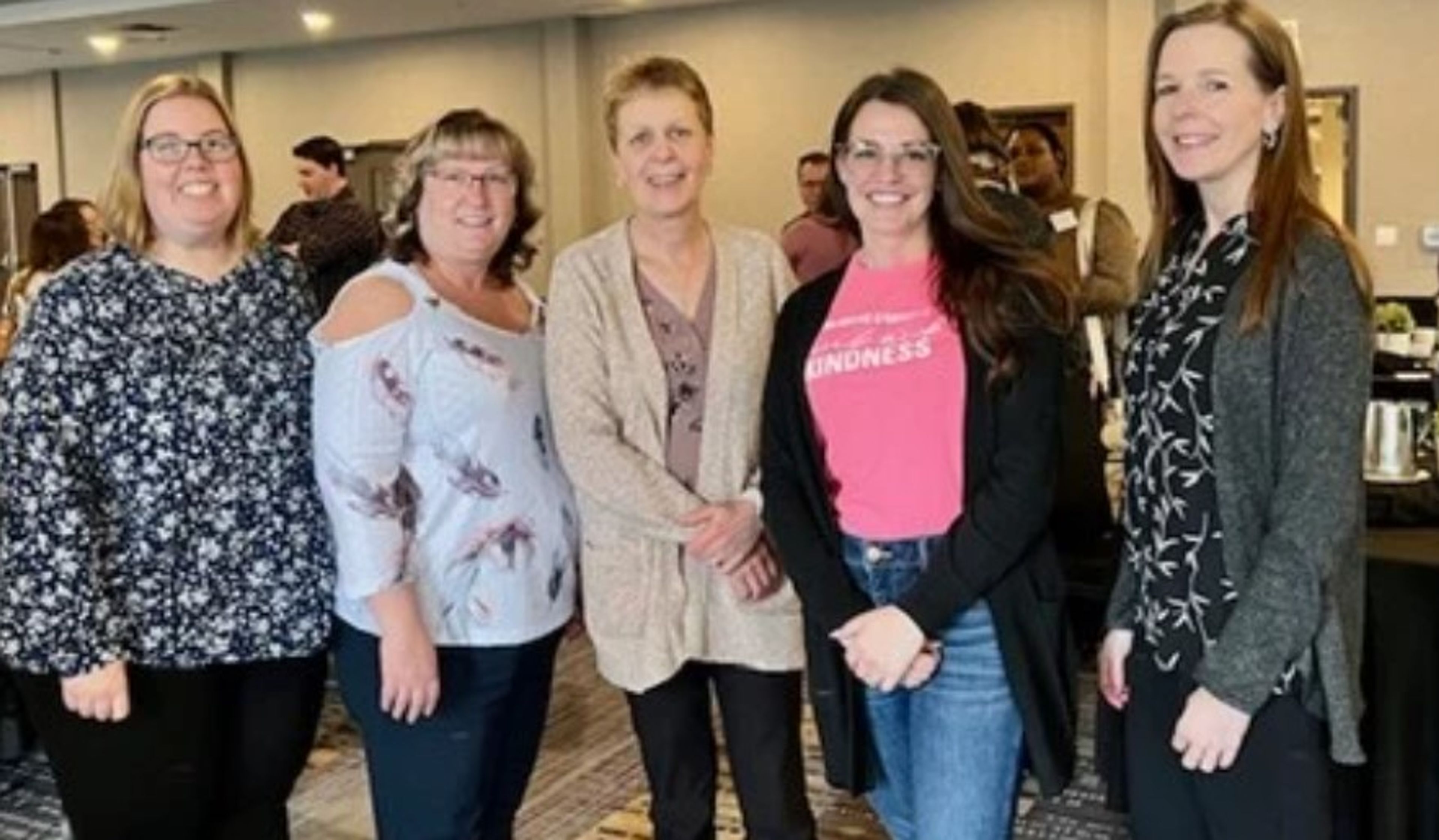 Five women stand together indoors, smiling for a group photo in a conference or meeting room. There are tables and people in the background.