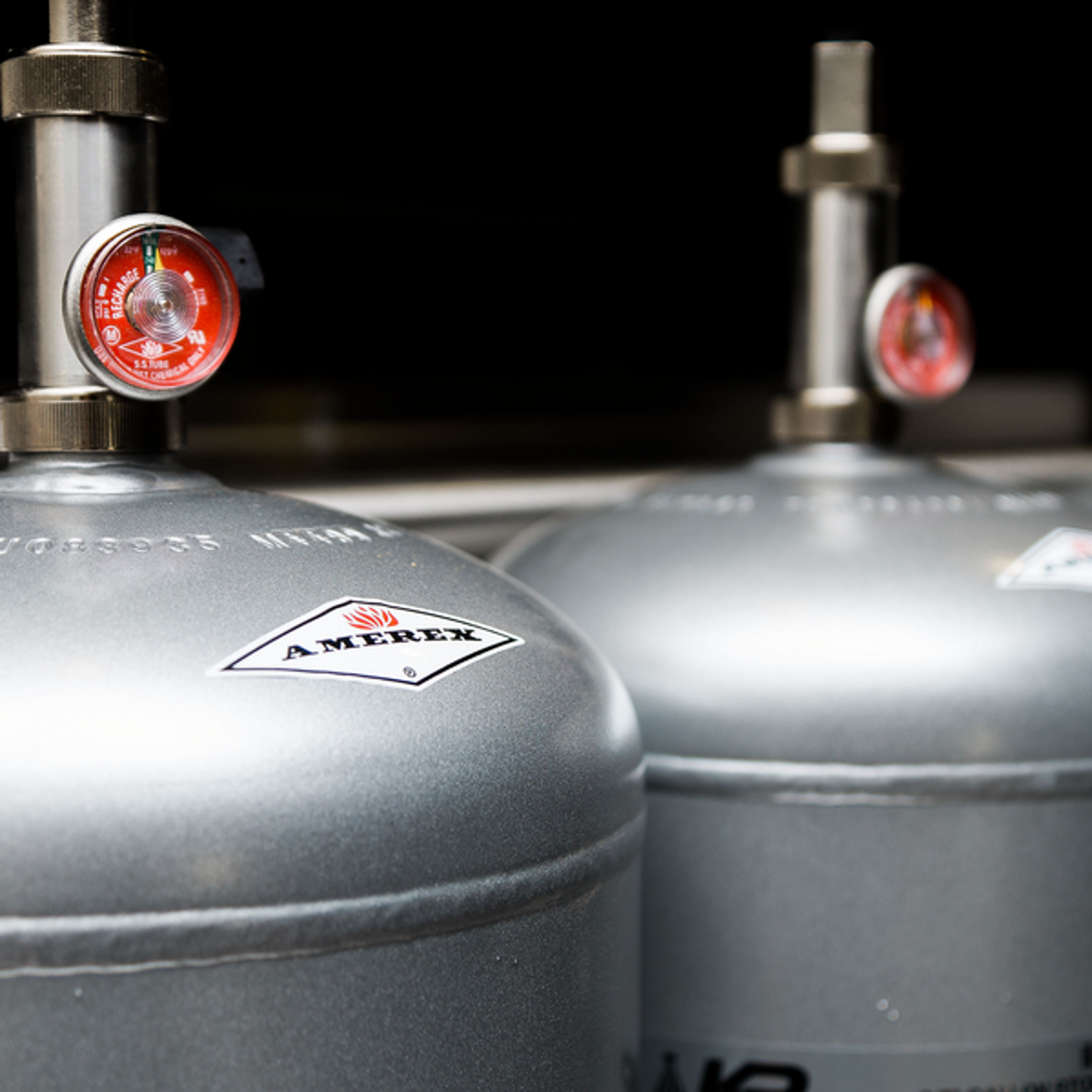 Close-up of two silver Amerex fire extinguisher tanks with pressure gauges, positioned side by side in an indoor setting.
