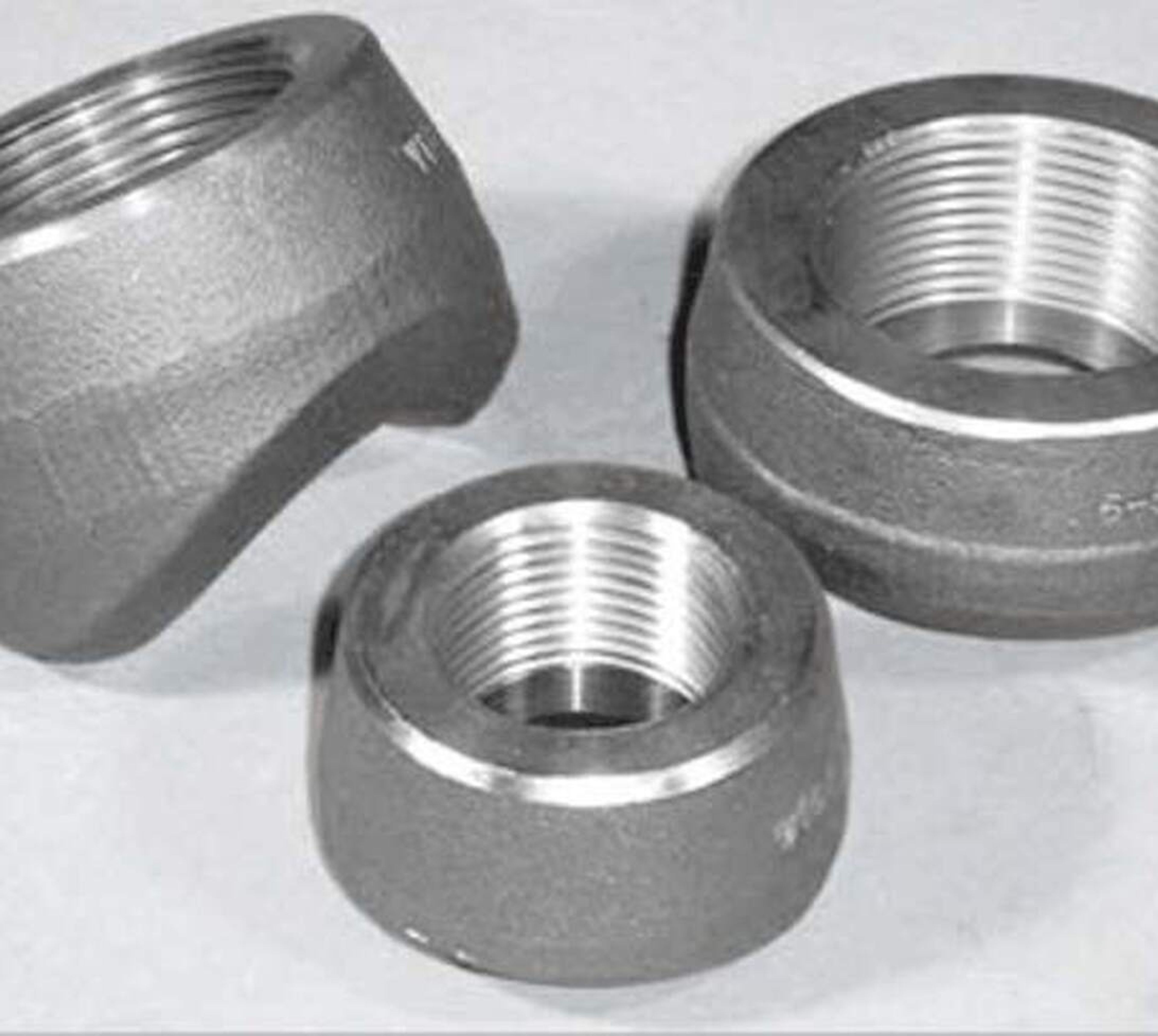 Three metallic pipe fittings with threaded interiors, placed on a light surface. Two are upright, showing the threading inside, while one fitting is tilted, revealing its shape and partial threads.