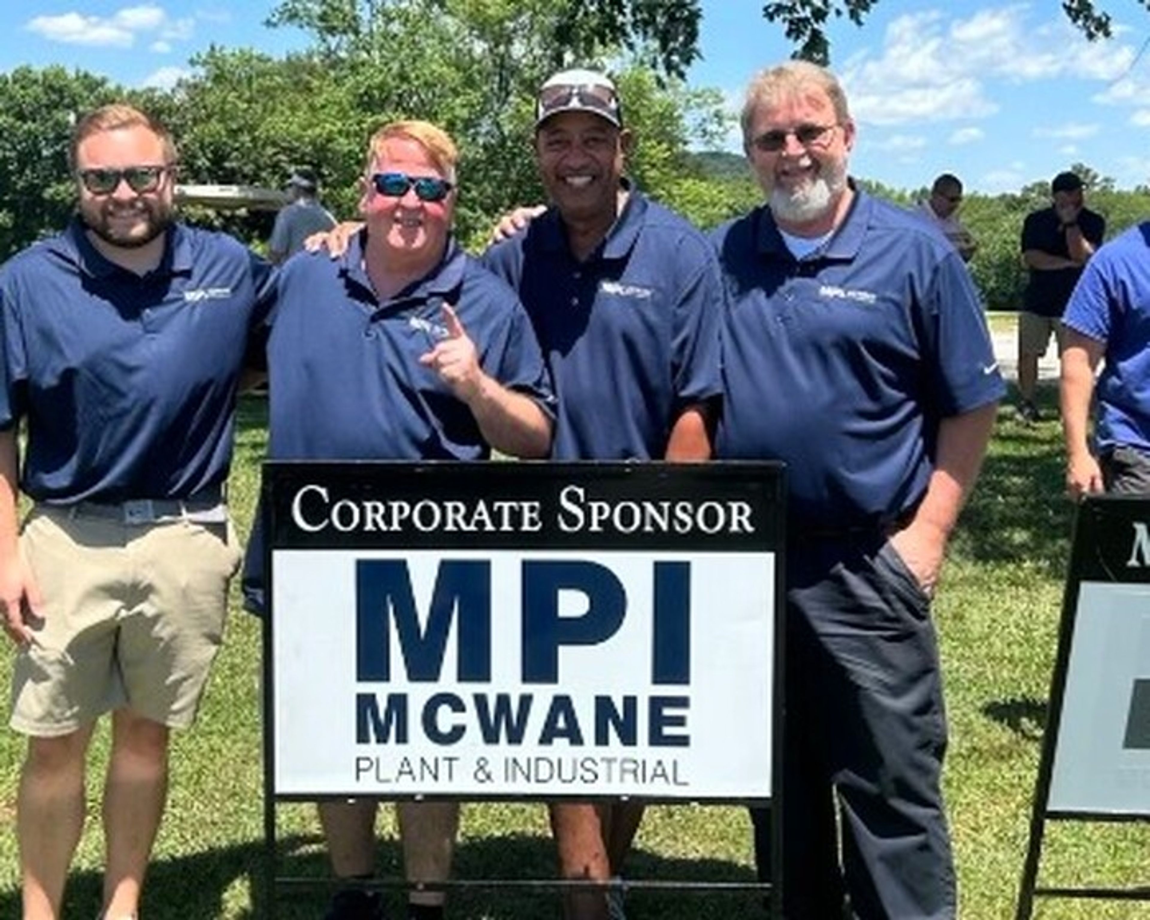 Four men in blue polo shirts stand outdoors on grass, smiling, with arms around each other behind a sign that reads “Corporate Sponsor MPI McWane Plant &amp; Industrial.” Trees and blue sky are visible in the background.