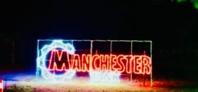 A neon light sign reads “MANCHESTER” in red letters, with a blue and white circular design on the left side, glowing at night.