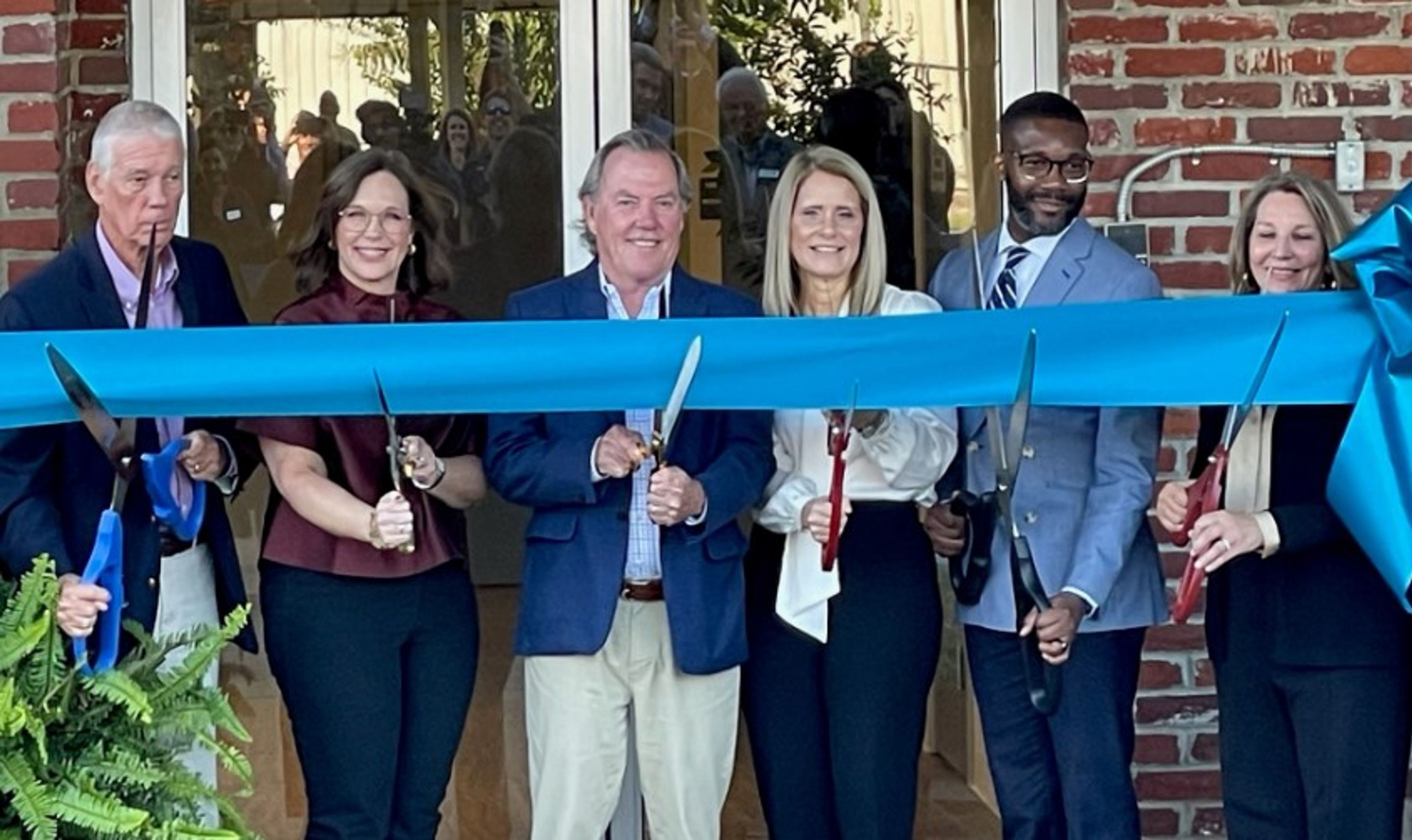 Six people stand in front of a building, smiling and holding scissors to cut a large blue ribbon during a ribbon-cutting ceremony. A brick wall and glass doors are visible in the background.