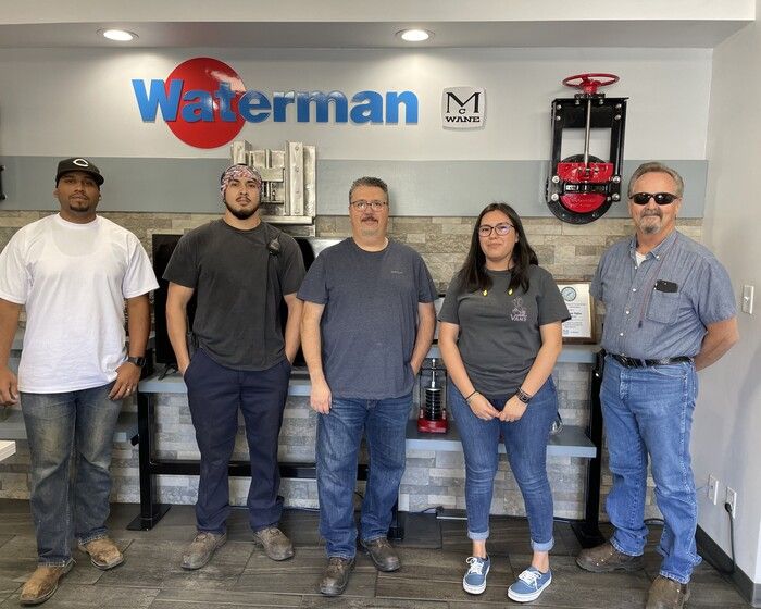 Five people stand in front of a Waterman company sign in an office, posing for a group photo. Equipment and metal tools are displayed on the wall behind them.