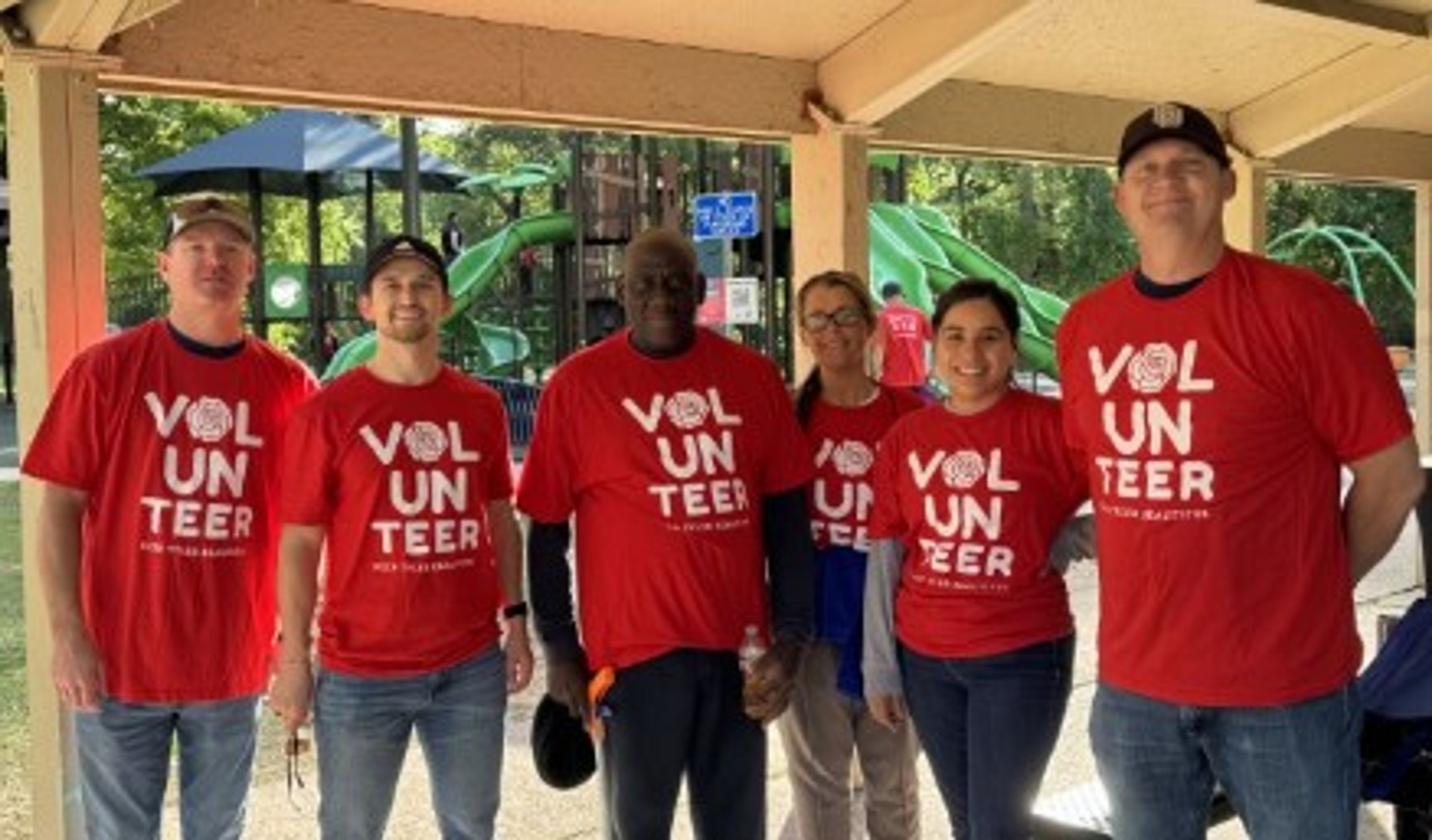 Six people wearing matching red “Volunteer” shirts stand together and smile under a covered outdoor area, with playground equipment visible in the background on a sunny day.