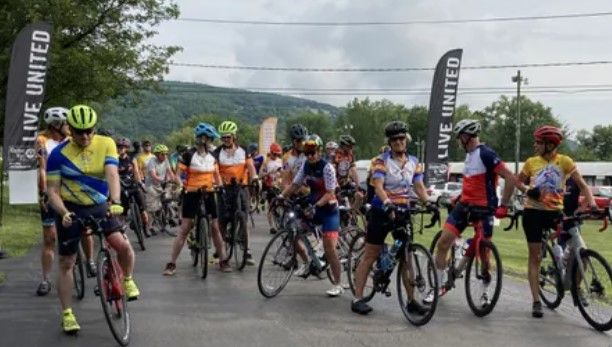 A group of cyclists in colorful jerseys and helmets gather on a road, preparing for a ride. Two black flags with “LIVE UNITED” text are visible, and trees and hills are in the background.