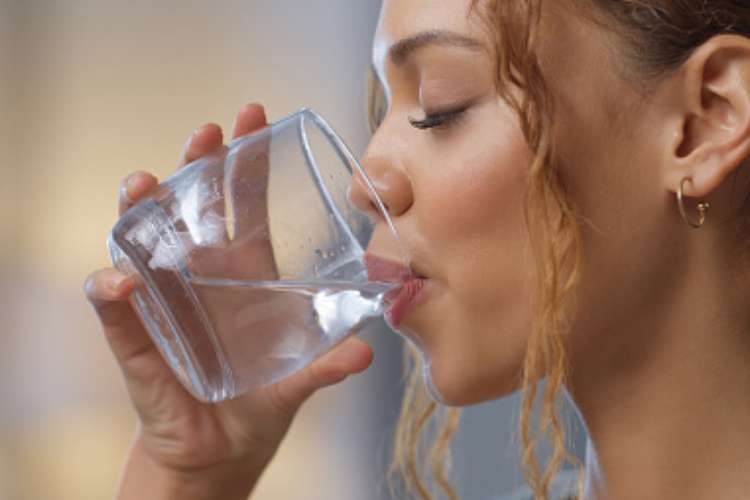 A woman with curly hair drinks a glass of water. She has a gold hoop earring and is shown in profile against a soft, blurred background.