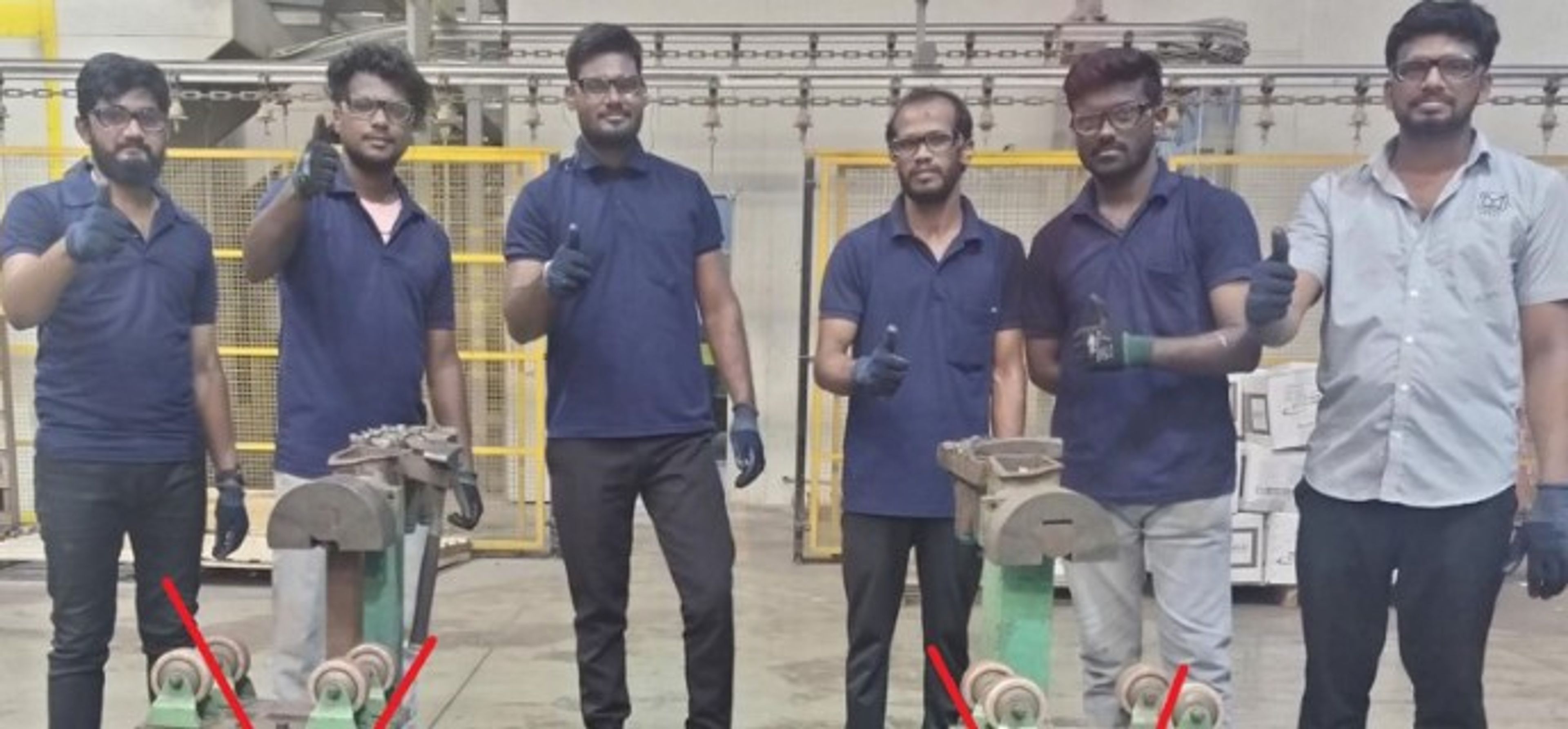 Five men wearing gloves pose in a workshop, each giving a thumbs up. Two metal machines are in front of them, each marked with a red X. Yellow safety barriers are visible in the background.