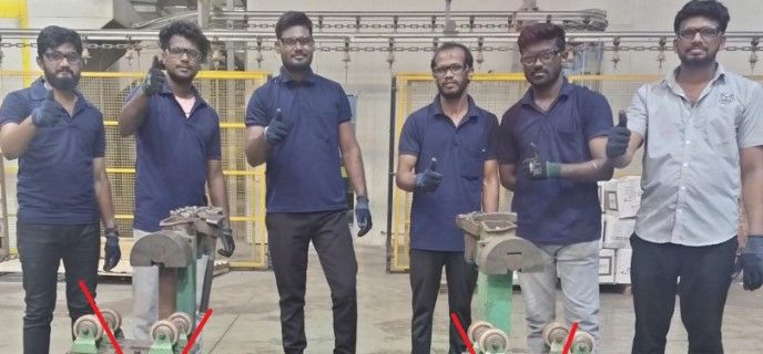 Five men wearing gloves pose in a workshop, each giving a thumbs up. Two metal machines are in front of them, each marked with a red X. Yellow safety barriers are visible in the background.