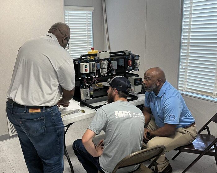 Three men are gathered around a table with technical equipment; two are seated while one stands, and they appear to be in discussion or training inside a small, bright room with a window.