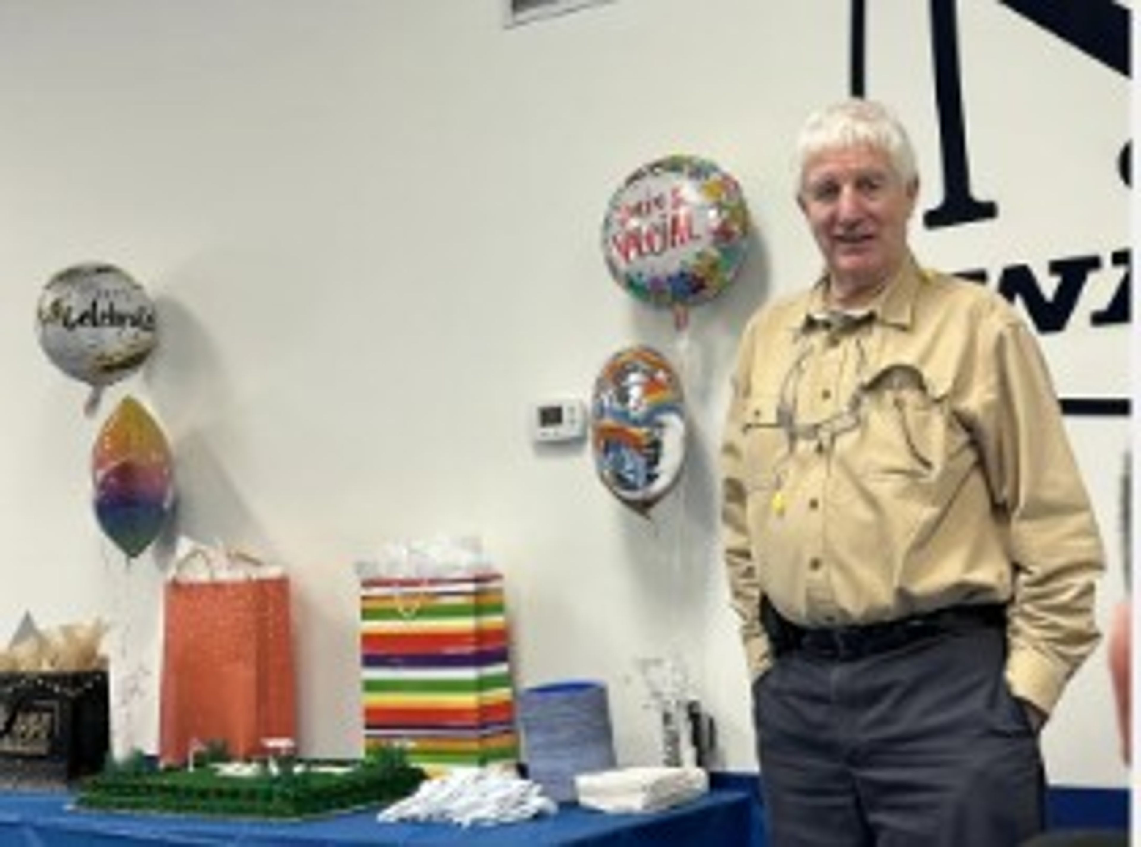 Older man in tan shirt stands beside a table with a cake, balloons, and colorful gift bags. The wall behind him has a large logo and celebration-themed decorations.