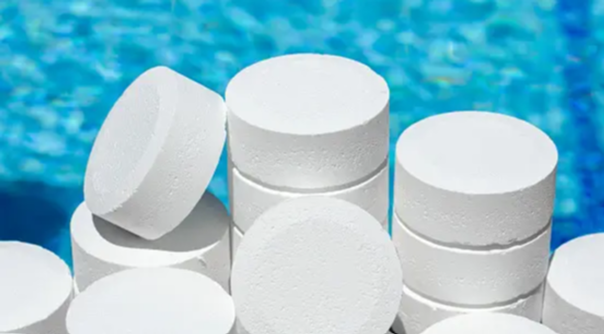A stack of large white chlorine tablets is shown in front of a blue swimming pool.