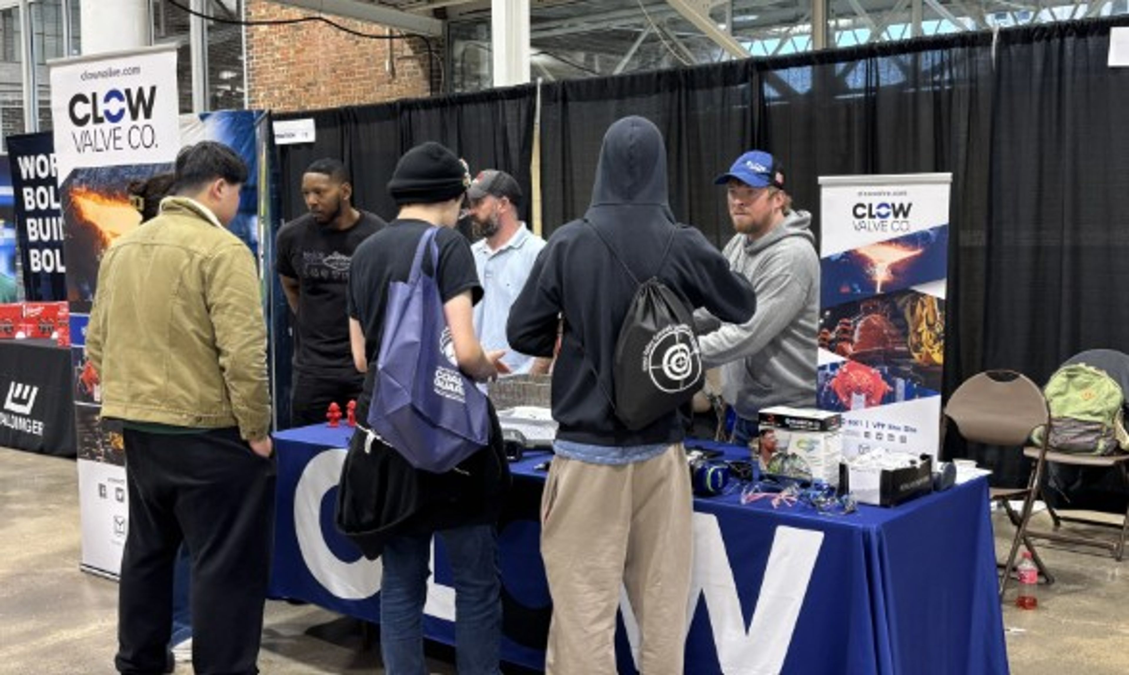 Five people stand at a booth with a “Clow Valve Co.” banner at an indoor event. Two men behind the table talk to three visitors. The table displays promotional items and a laptop.