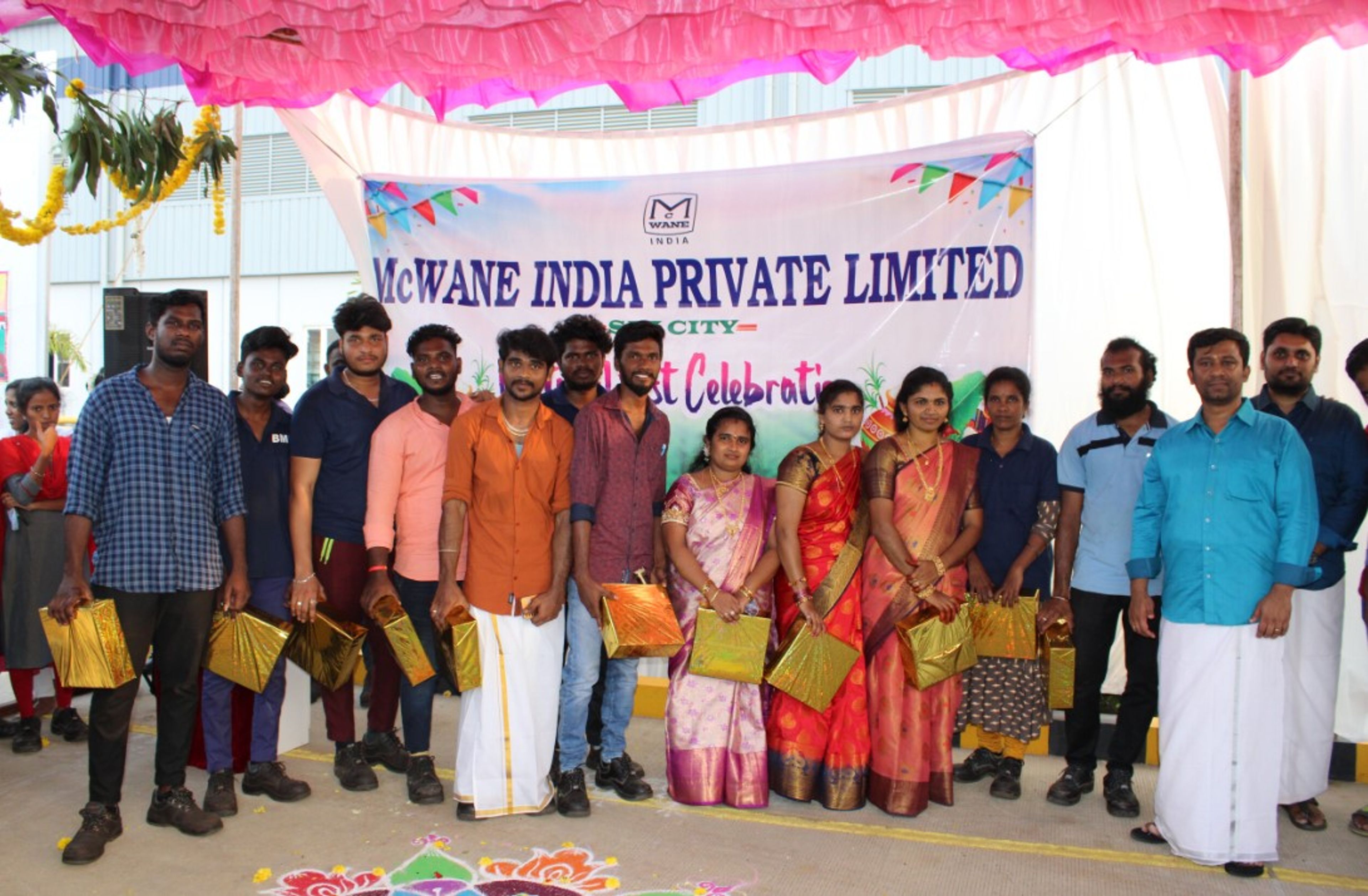 A group of people stand in front of a banner reading “McWane India Private Limited,” holding gold gift bags, dressed in festive attire, possibly celebrating a company event or festival.