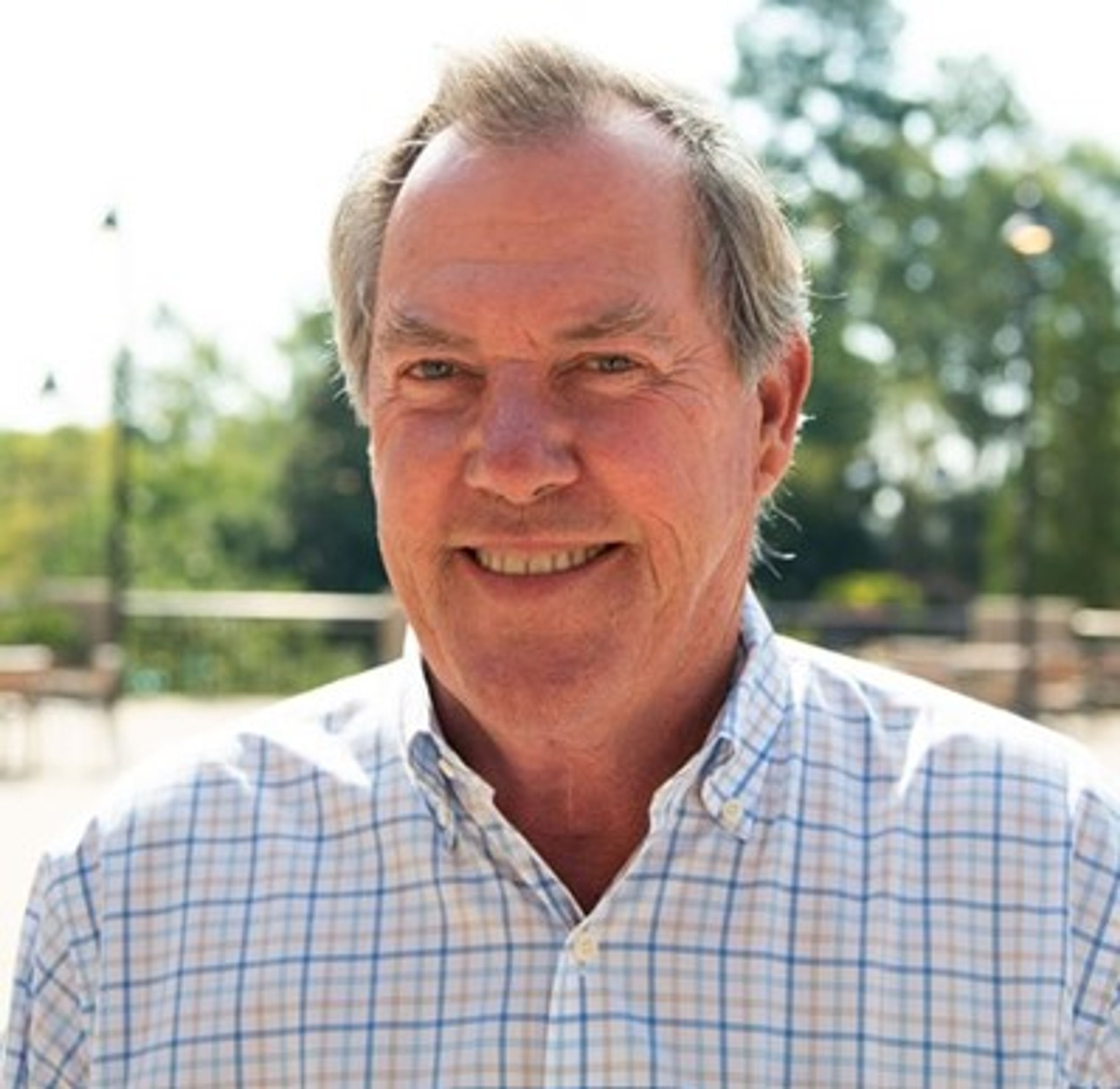 Smiling older man with short gray hair wearing a light-colored plaid shirt, standing outdoors on a sunny day with trees and a blurred background behind him.