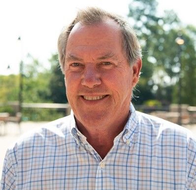 Smiling older man with short gray hair wearing a light-colored plaid shirt, standing outdoors on a sunny day with trees and a blurred background behind him.