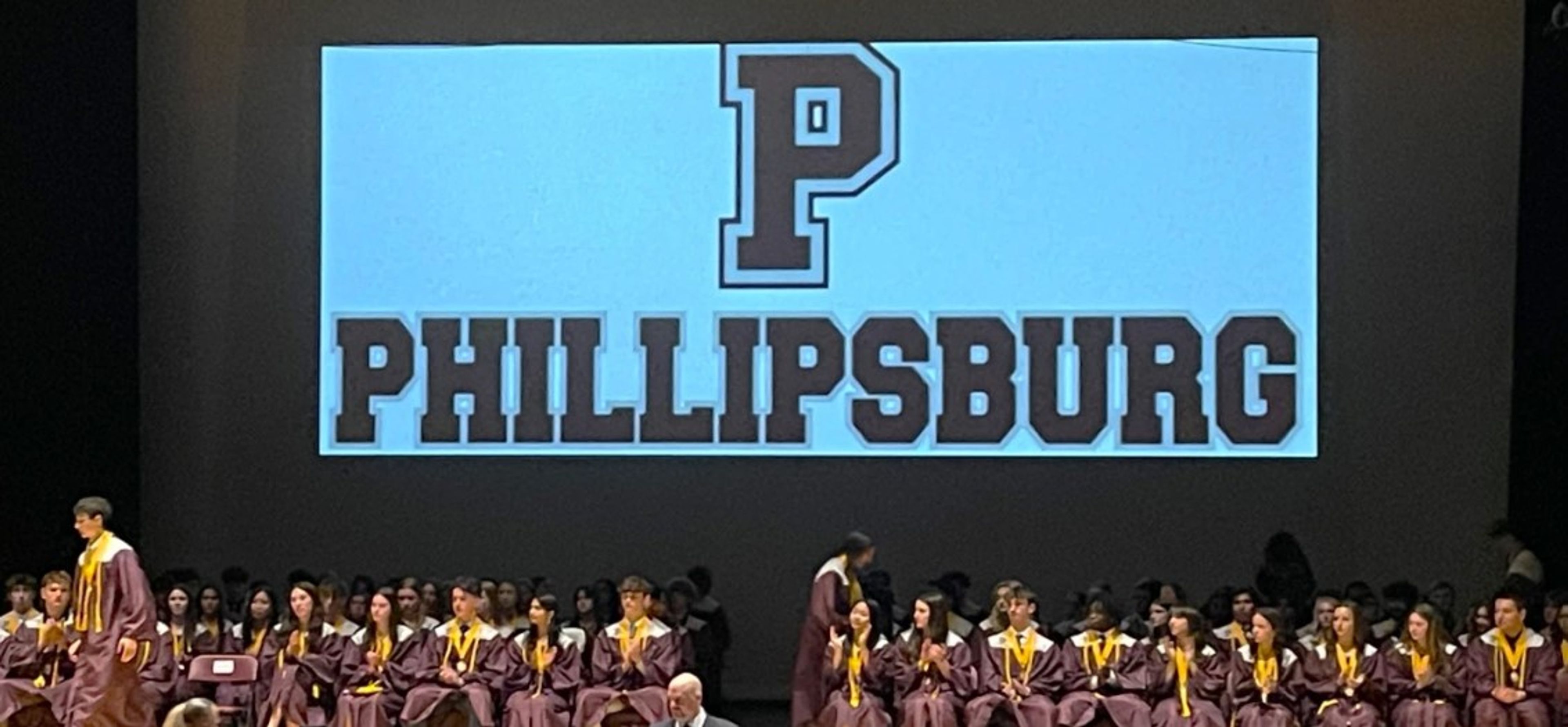 Graduates in maroon gowns sit on stage with a large screen behind them displaying a big letter “P” and the word “PHILLIPSBURG” in bold letters.