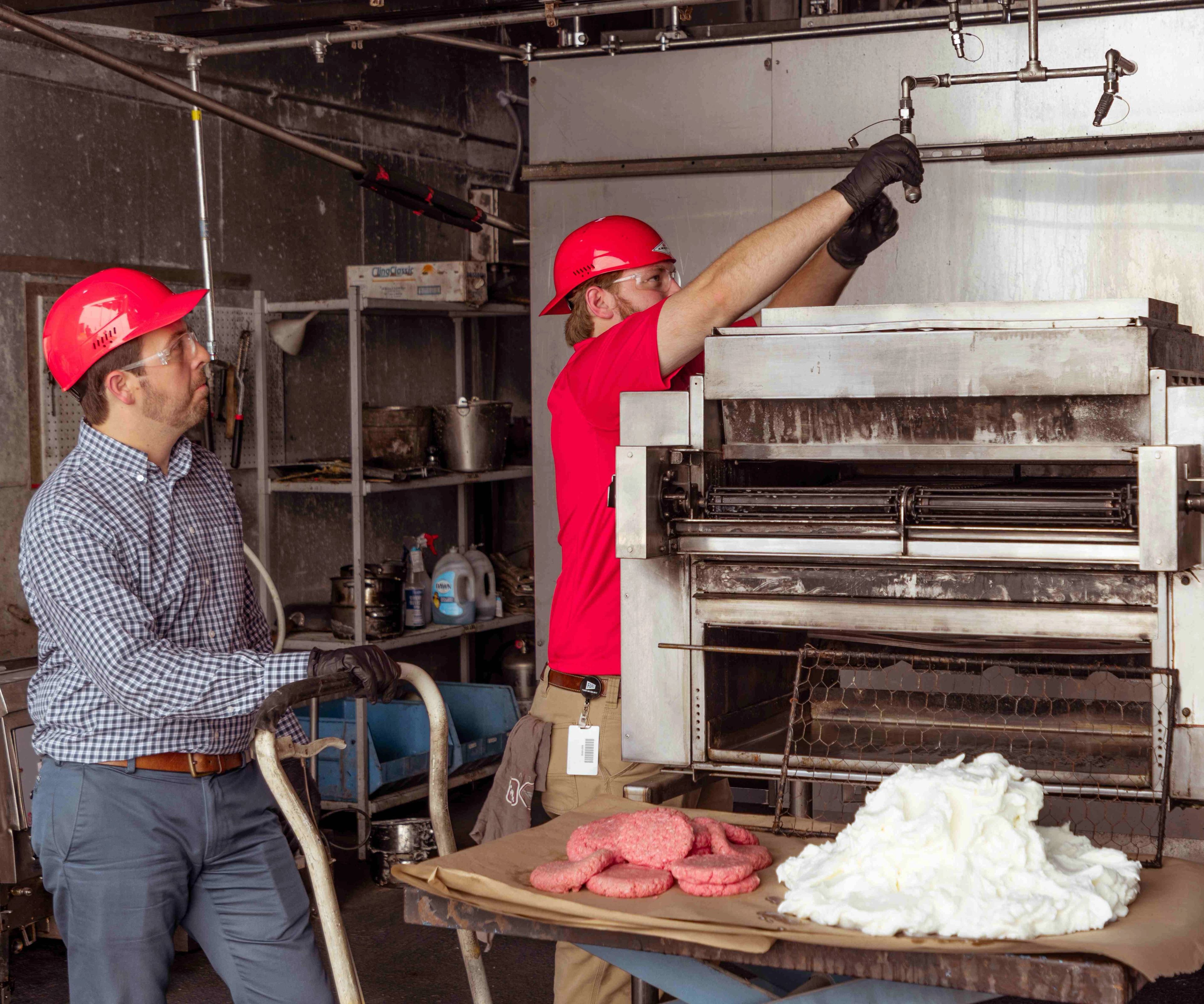 Two people in safety gear work in an industrial kitchen, one adjusting equipment while the other observes. Raw burger patties and a pile of white substance sit on a tray in the foreground.