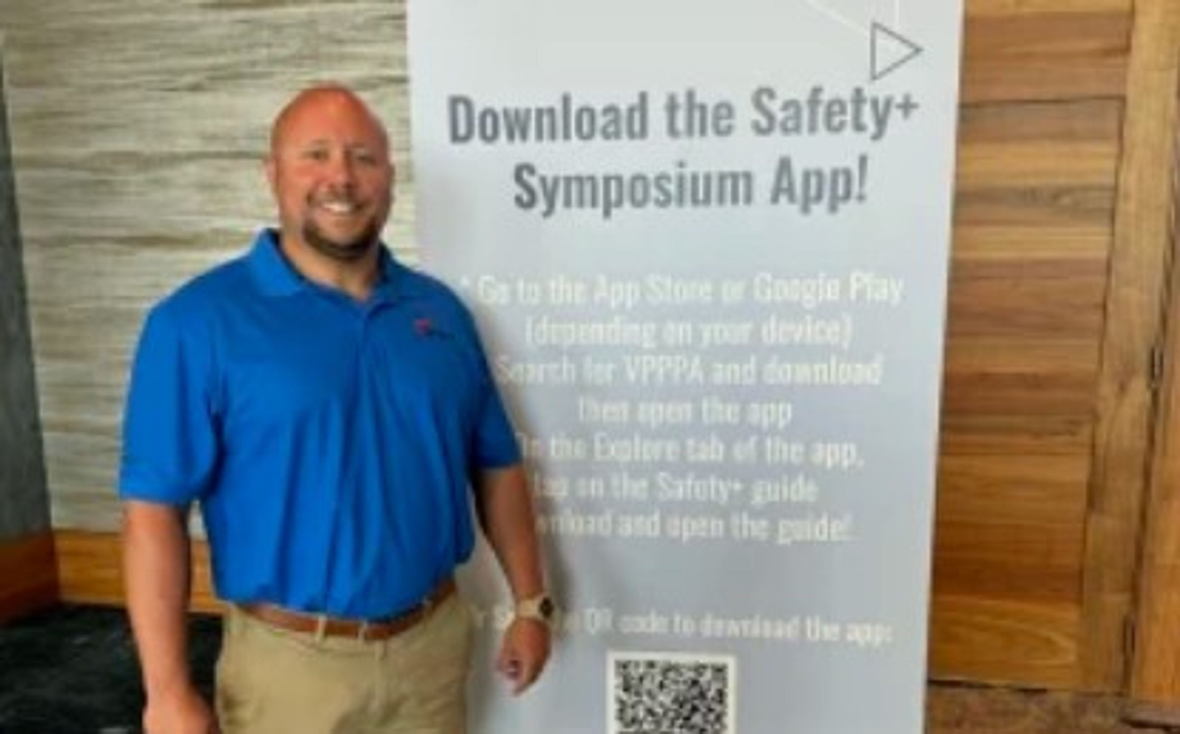 A man in a blue polo shirt stands next to a large sign promoting the Safety+ Symposium app, with app download instructions and a QR code visible on the sign.