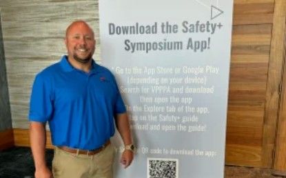 A man in a blue polo shirt stands next to a large sign promoting the Safety+ Symposium app, with app download instructions and a QR code visible on the sign.