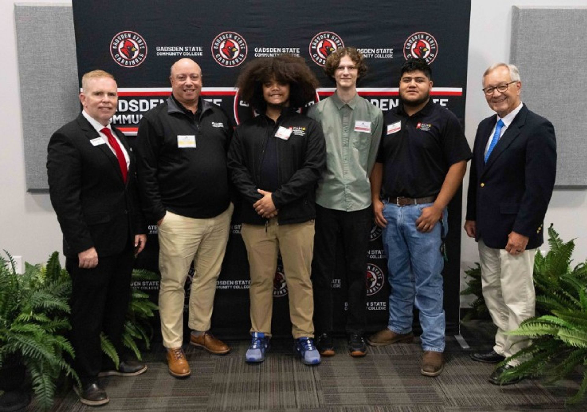 Six men stand in front of a Gadsden State Community College backdrop, smiling for a group photo. Four men are wearing slacks and dress shirts, and two are in business suits. Ferns are visible at the bottom of the image.