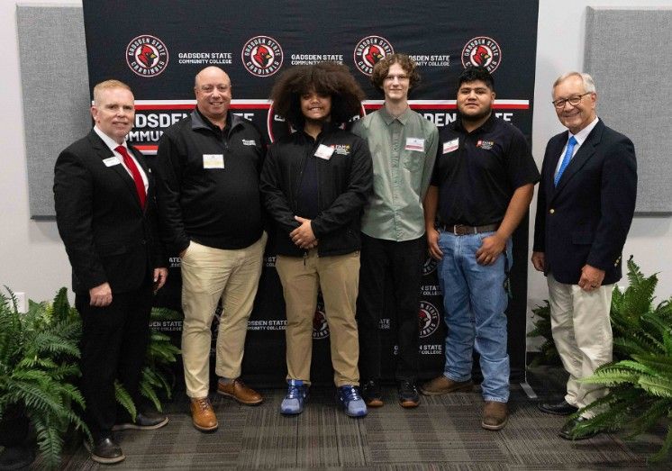 Six men stand in front of a Gadsden State Community College backdrop, smiling for a group photo. Four men are wearing slacks and dress shirts, and two are in business suits. Ferns are visible at the bottom of the image.