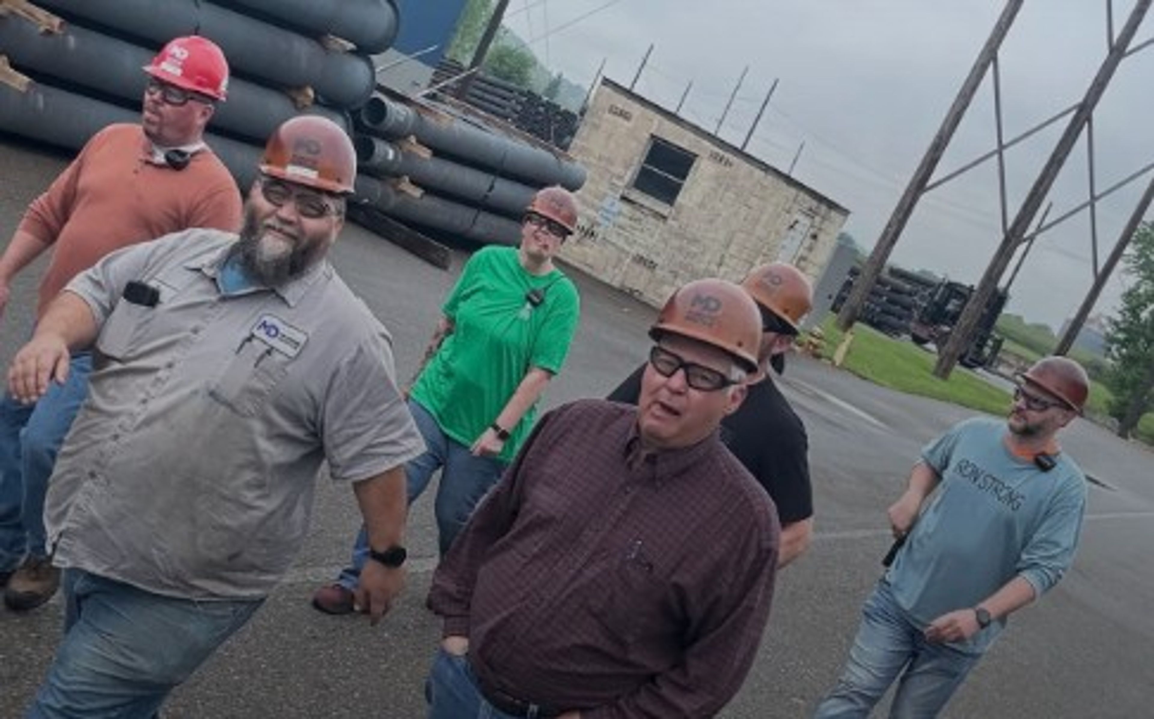 A group of six people wearing hard hats and casual clothing walk outside near industrial pipes and a small building on a cloudy day.