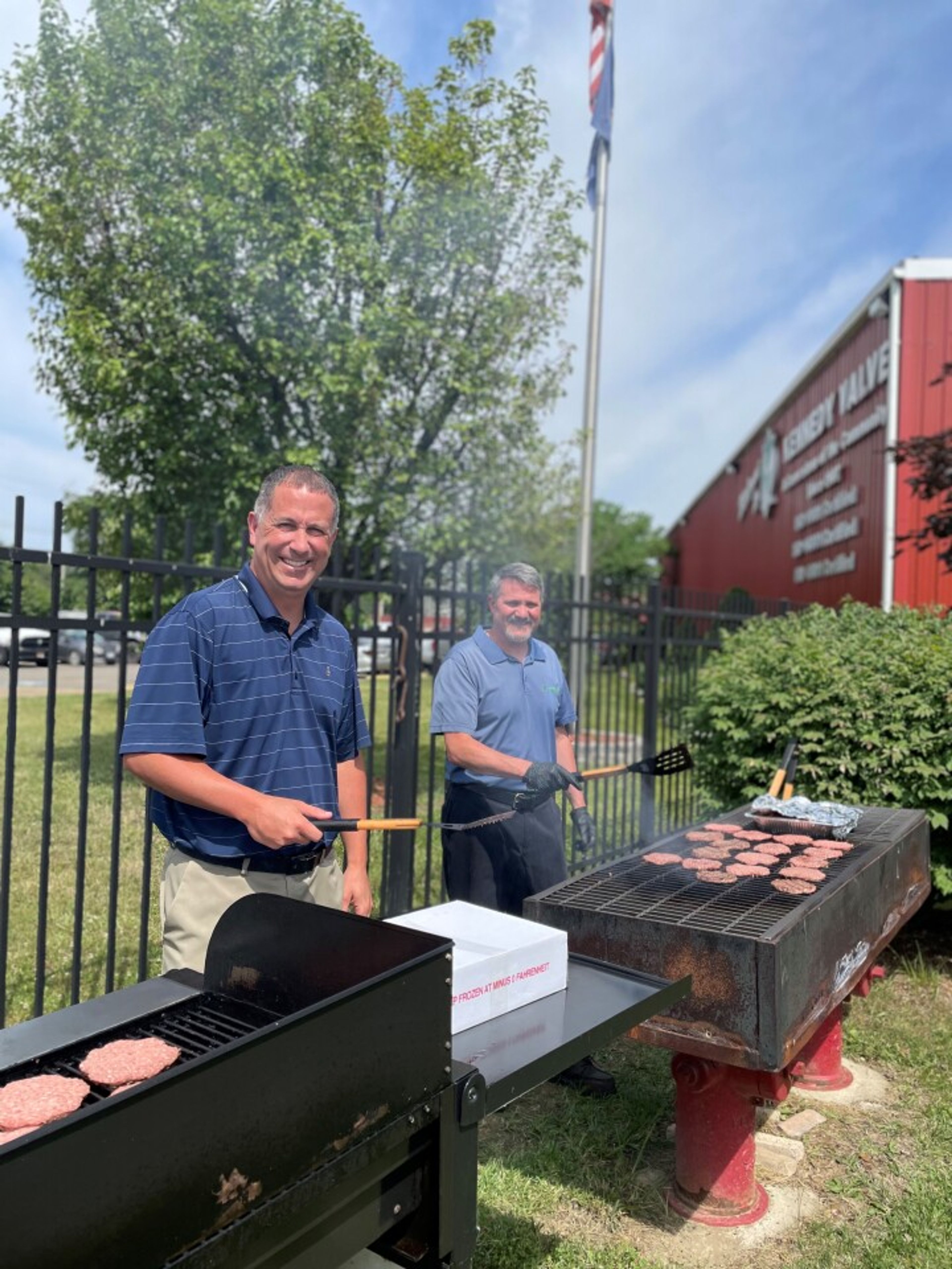 Two men grill burgers outdoors on a sunny day, standing by metal grills with spatulas in hand. A red building, green trees, and an American flag are visible in the background.