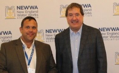 Two men in business attire stand and smile in front of a backdrop with the NEWWA (New England Water Works Association) logo repeatedly printed on it.
