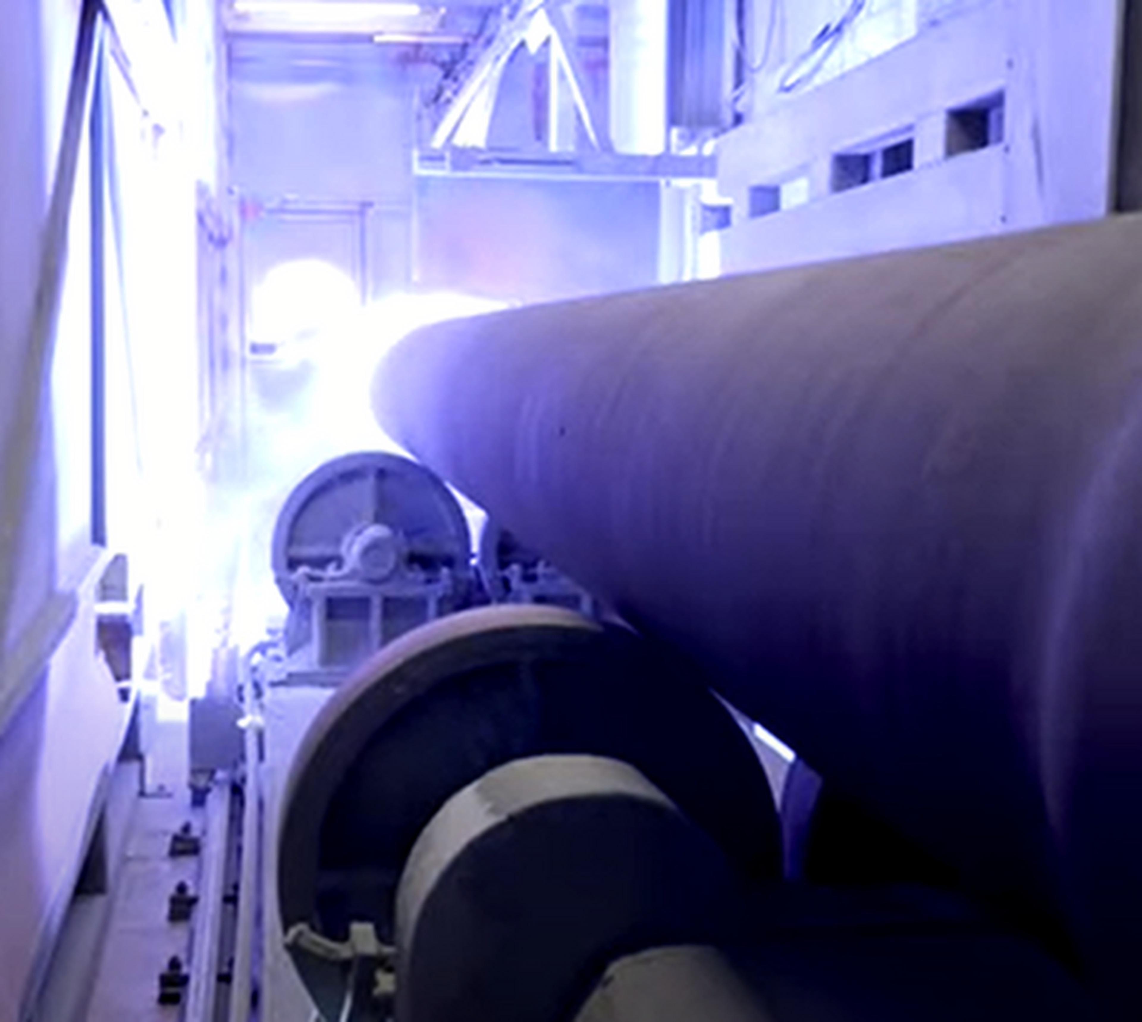 Large cylindrical pipes move through an industrial facility, illuminated by bright blue-white lights, likely representing a high-temperature or welding process. The scene has a modern, technical atmosphere.