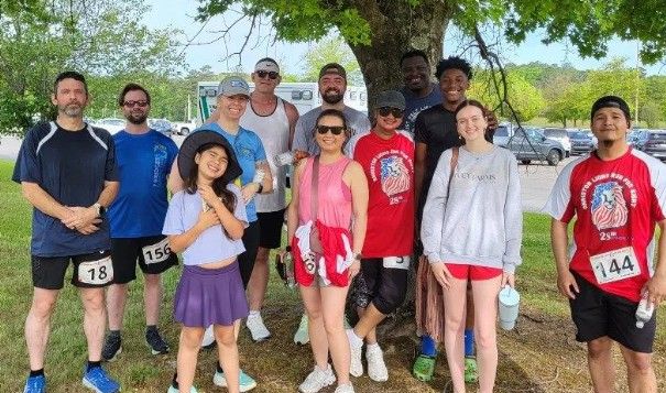 A diverse group of twelve people, some in athletic wear with race bibs, pose and smile together under a large tree on a grassy area, suggesting a fun run or race event.