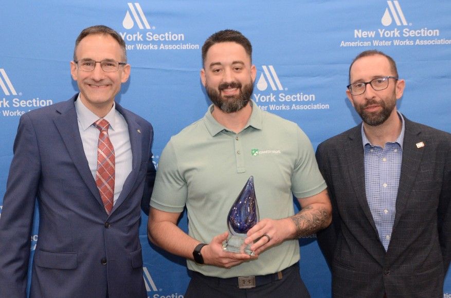Three men stand in front of a New York Section American Water Works Association backdrop. The man in the center holds a blue glass award and smiles, while the other two stand on either side, also smiling.