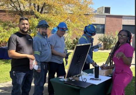 Five people stand outdoors at a table; three wear blue work uniforms and helmets, while two are in casual clothes. A woman in pink stands beside the table with paperwork and a sign, with a brick building in the background.