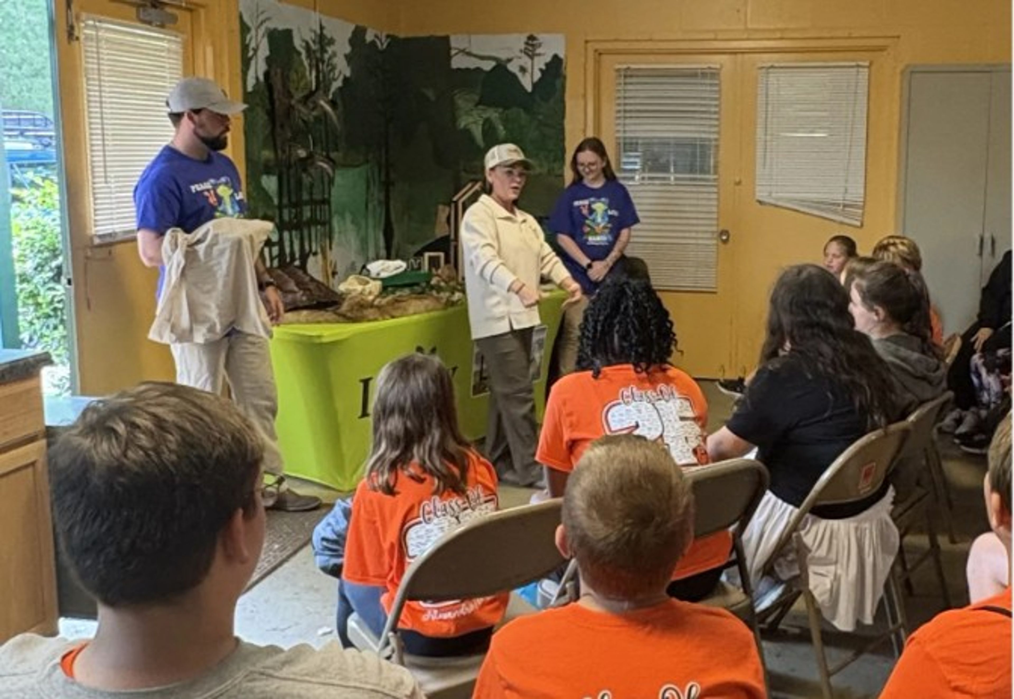 A group of children in orange shirts sits on chairs, listening to a woman speak at the front of a classroom. Two adults in purple shirts stand nearby. There are nature-themed decorations and animal displays in the background.