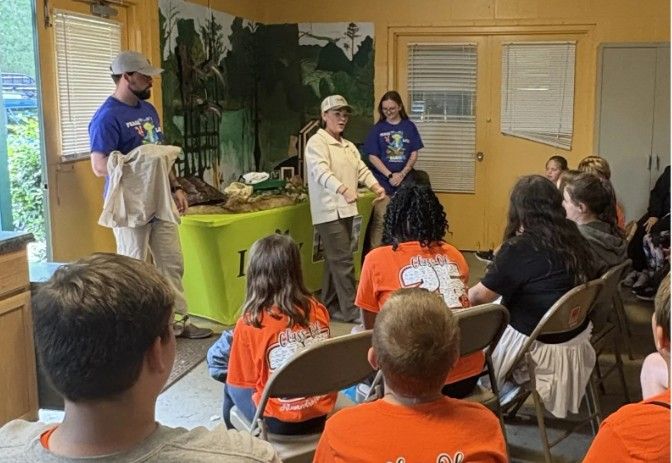 A group of children in orange shirts sits on chairs, listening to a woman speak at the front of a classroom. Two adults in purple shirts stand nearby. There are nature-themed decorations and animal displays in the background.