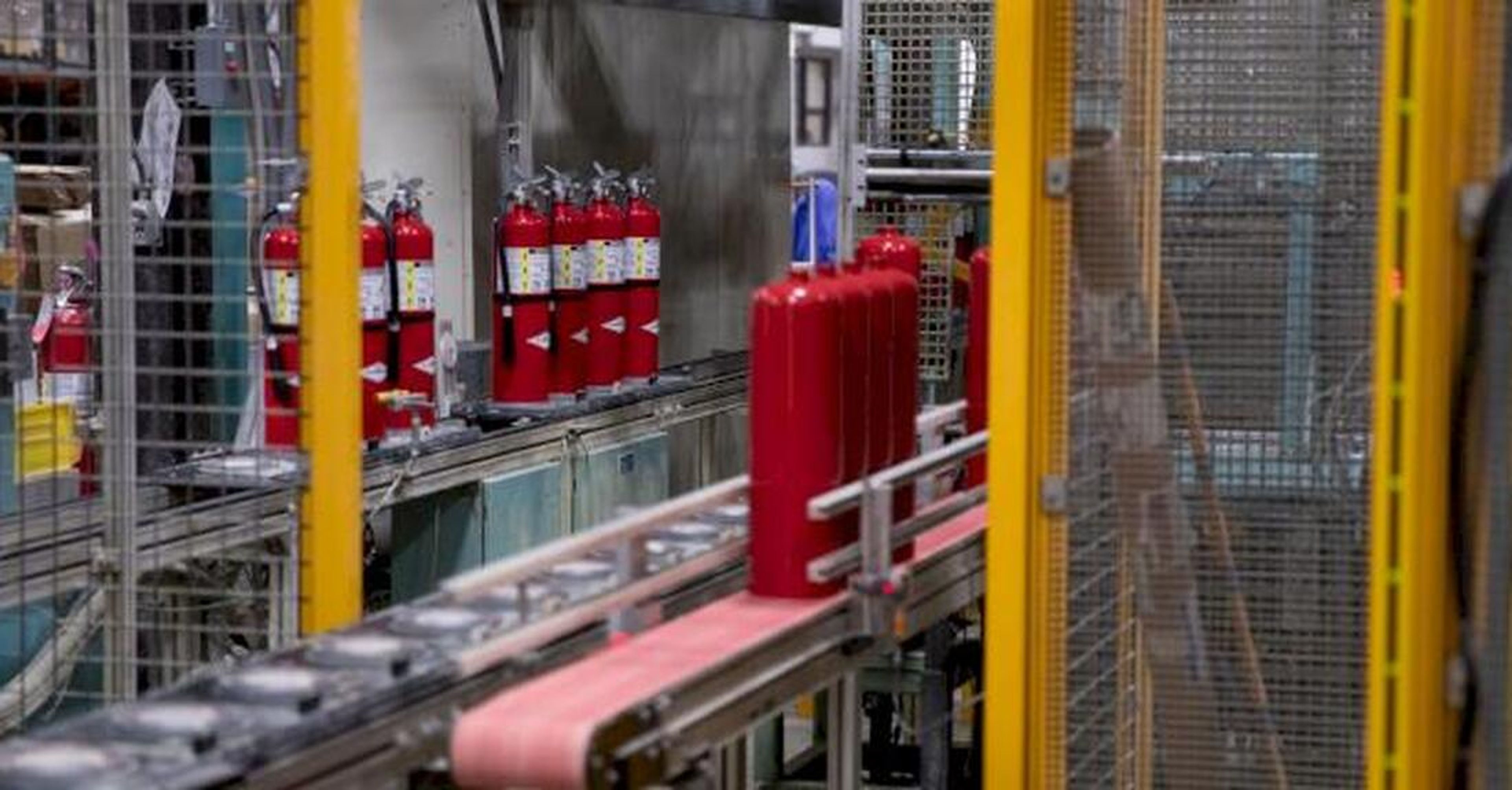 Red fire extinguishers move along a conveyor belt inside a factory, surrounded by metal fencing and machinery.