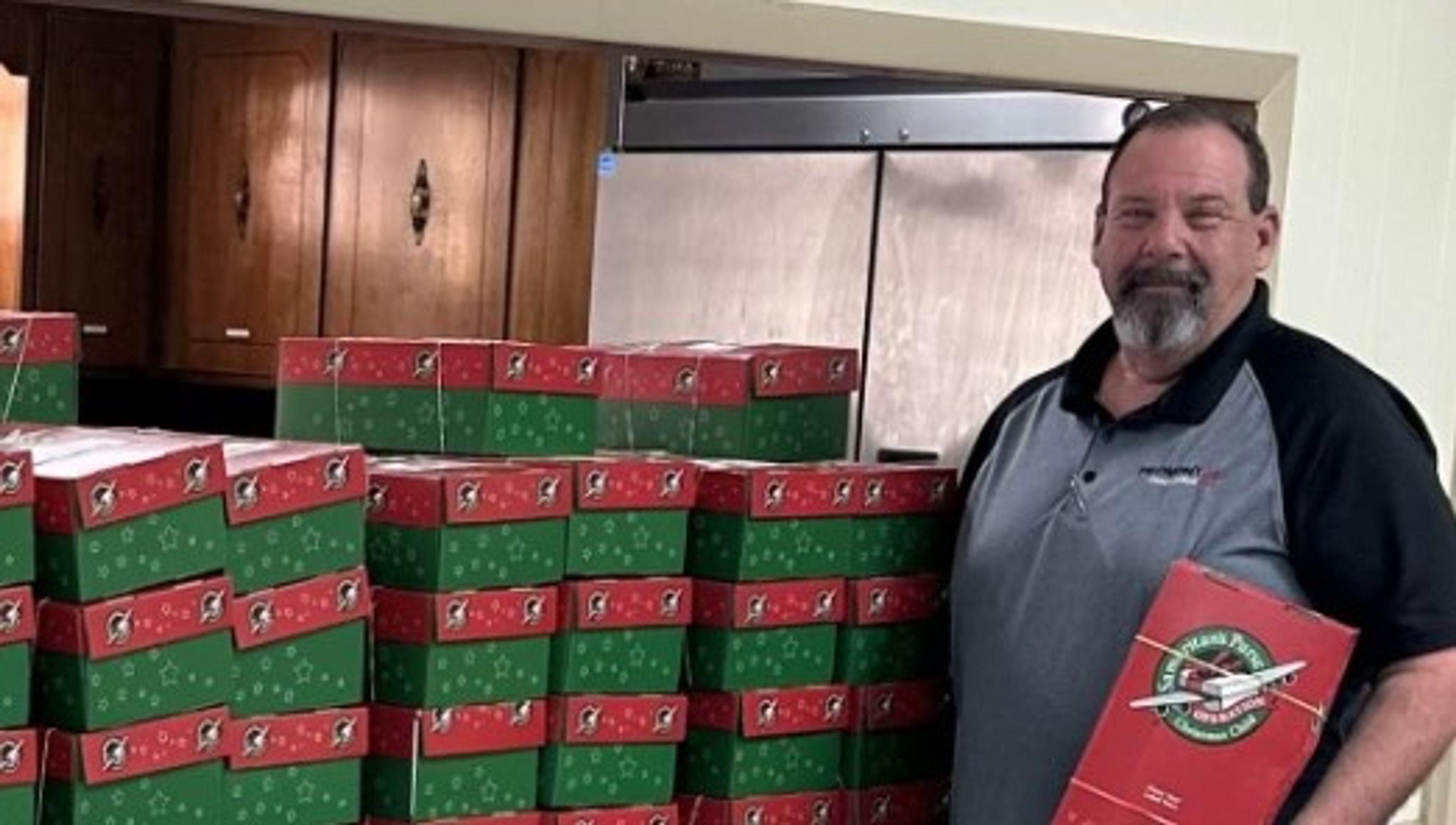 A man stands beside a stack of red and green shoeboxes, holding one in his hand. He is indoors, smiling, and there are cabinets and a refrigerator in the background.