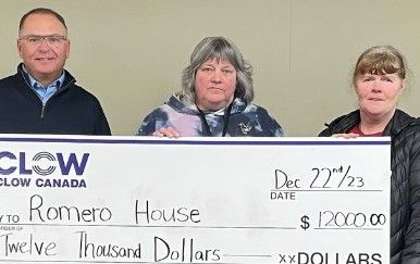 Three people stand holding a large check for $12,000 made out to Romero House from Clow Canada, dated December 22, 2023. The background is plain and indoor.