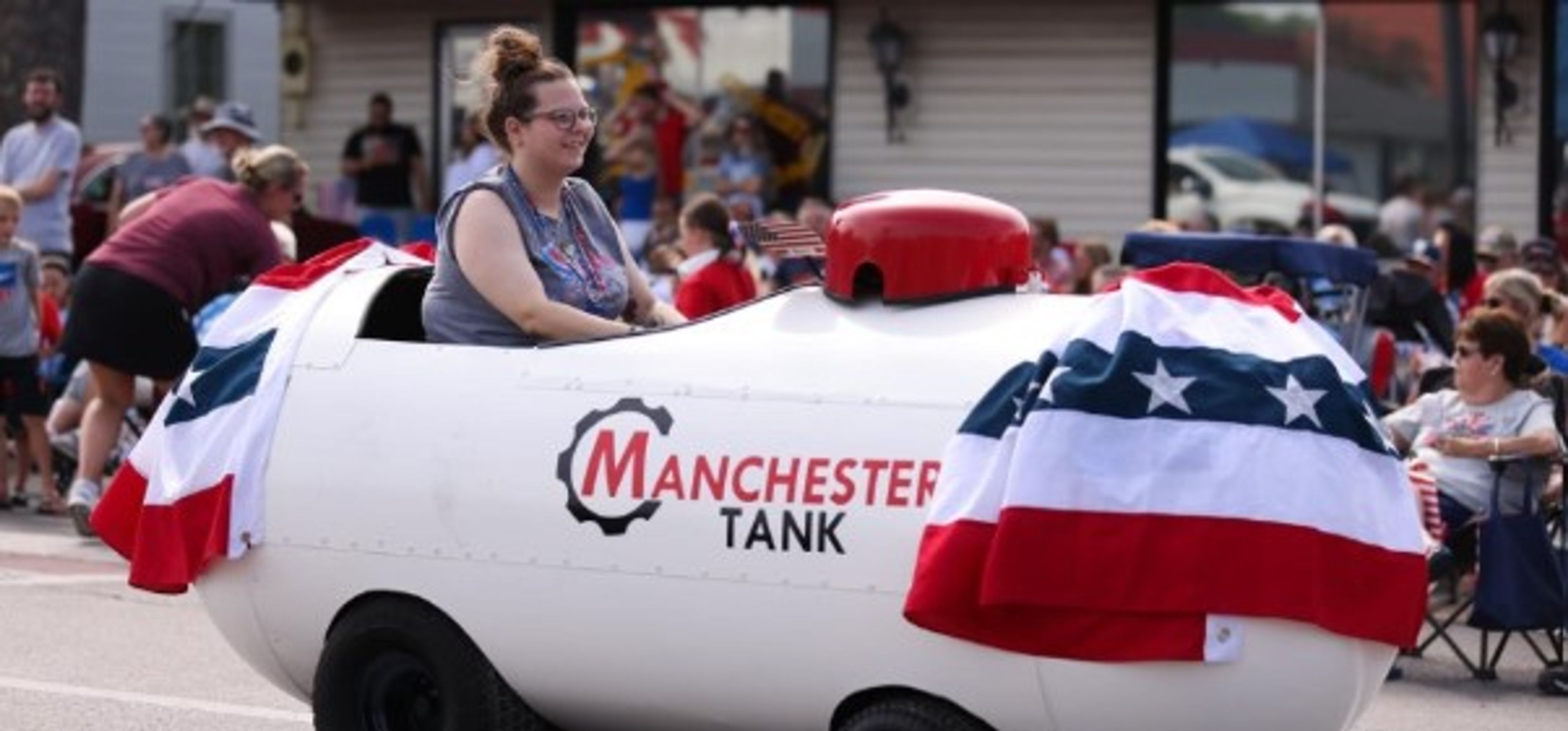 A woman rides a propane tank-shaped vehicle decorated with patriotic bunting in a parade. “Manchester Tank” is printed on the side, and people watch from the background.
