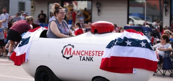 A woman rides a propane tank-shaped vehicle decorated with patriotic bunting in a parade. “Manchester Tank” is printed on the side, and people watch from the background.