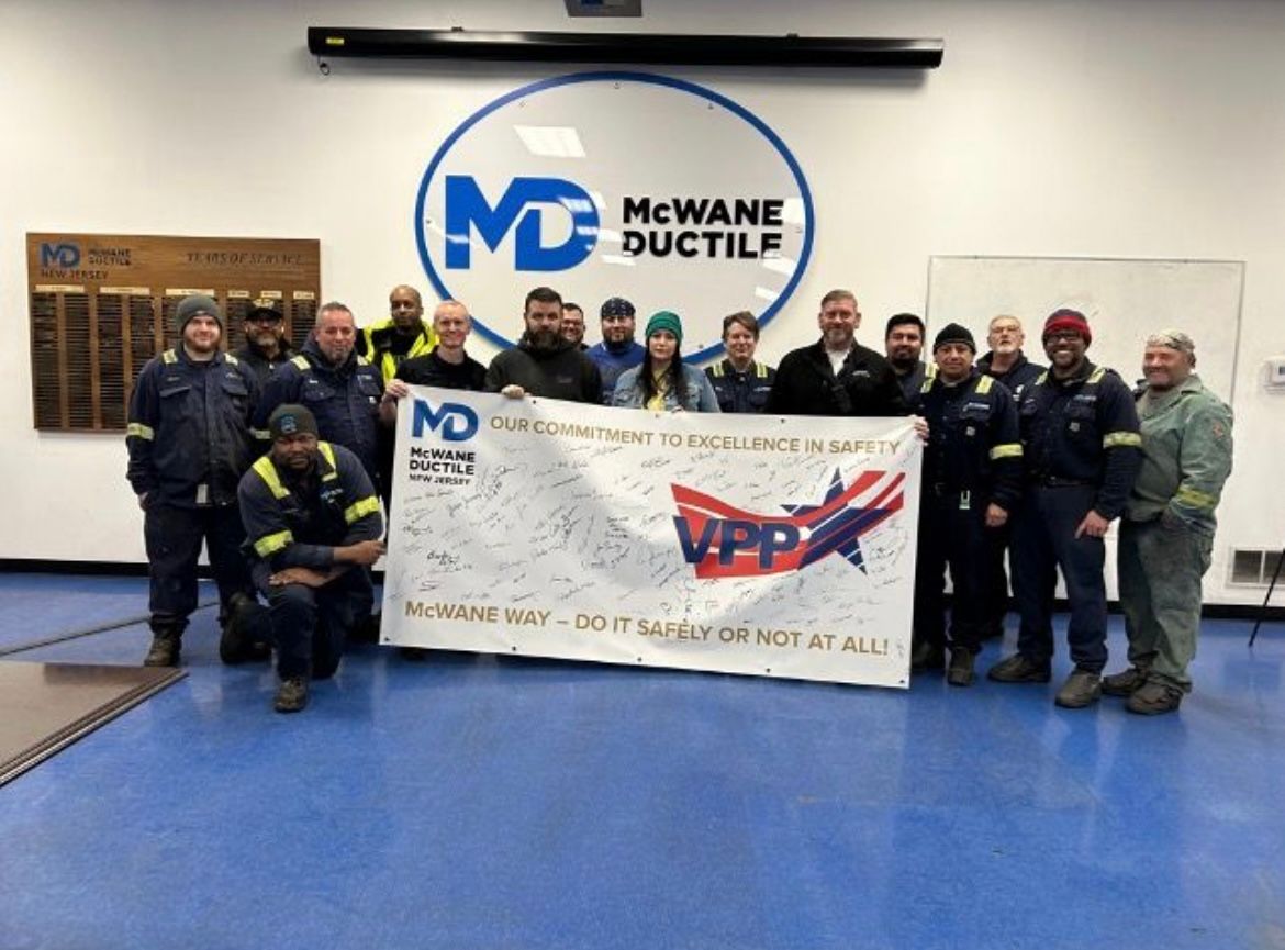 Group of workers in uniform and winter hats stand indoors holding a large safety banner with signatures; the McWane Ductile logo is visible on the wall behind them.